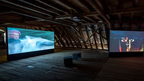In einem Dachraum aus Holz stehen zwei große Bildschirme, auf denen Filme laufen. 