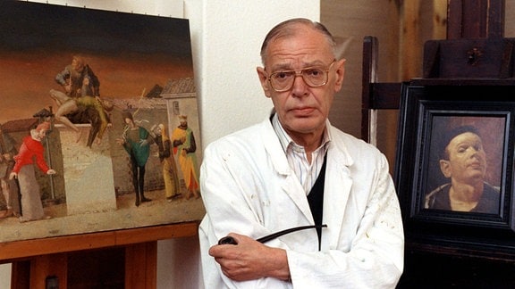 Der Leipziger Maler und Grafiker Werner Tübke: Ein älterer Mann mit kurzem, gescheiteltem grauen Haar und Brille steht in einem weißen Kittel zwischen zwei Gemälden. 