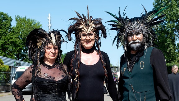 Besucher des WGT 2023: Zwei Frauen und ein Mann, die überwiegend in Schwarz gekleidet sind und aufwendigen Kopfschmuck tragen.