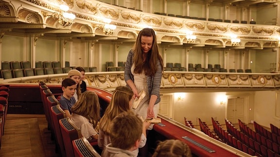 In der Semperoper sitzen mehrere Kinder auf roten Samtstühlen und schauen Balletschuhe an, die ihnen eine Frau zeigt. 