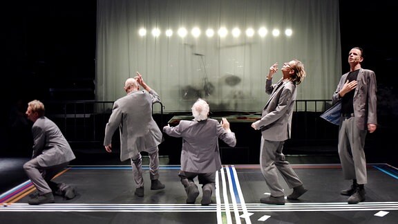  Fünf Männer in grauen Anzügen tanzen auf einer Bühne. Hinter einem milchig-durchsichtigen Vorhang ist ein Schlagzeug zu erkennen.