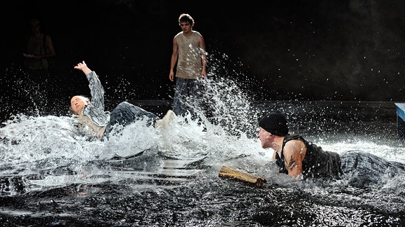 Auf einer Bühne, die unter Wasser steht, werfen sich zwei Personen auf den Boden, dass das Wasser spritzt.