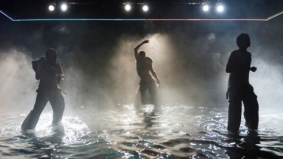 Drei Silhouetten tanzen auf einer Bühne. Das Licht kommt von hinten und spiegelt sich in dem Wasser auf dem Boden.