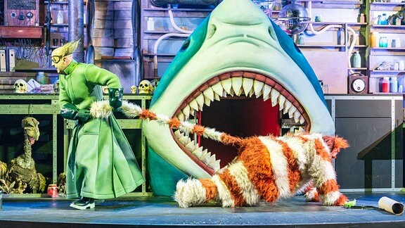 Eine Person in grünem Mantel zieht in eine Person in gestreiften Katzenkostüm über die Bühne, die wie ein verrücktes Labor eingerichtet ist.