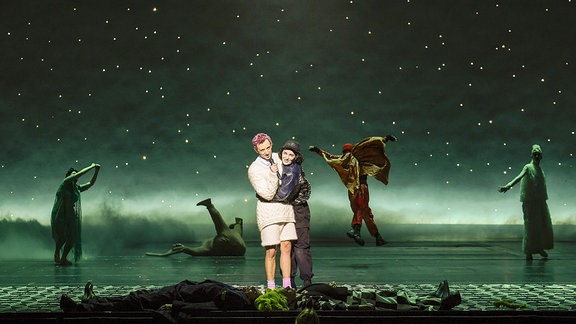 Zwei Personen stehen auf einer Bühne und klammern sich aneinander. Im Hintergrund bewegen sich Gestalten unter einem Sternenhimmel.