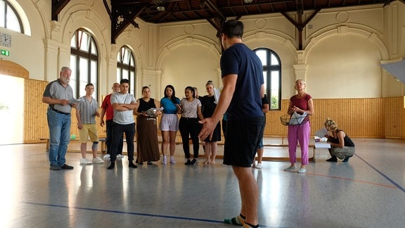 Eine Gruppe von Menschen steht auf einer improvisierten Theaterbühne in einer alten Turnhalle.