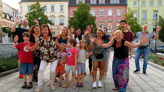 Ein mittelgroße Menschengruppe steht auf einem Platz und winkt in die Kamera.