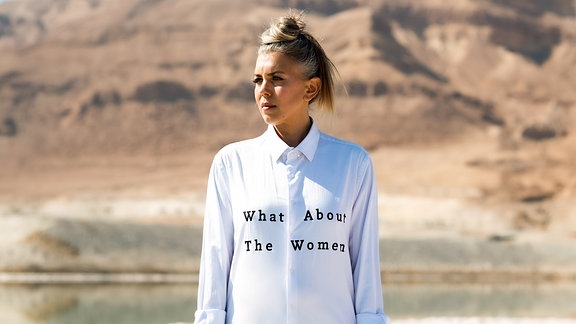 Ein Frau steht in einer Wüste. Auf ihrem weißen Hemd steht "What about the woman".