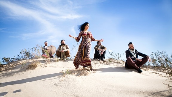 Yamma, eine Frau in einem langen Kleid steht auf einer Düne, im Hintergrund sitzen vier Männer.