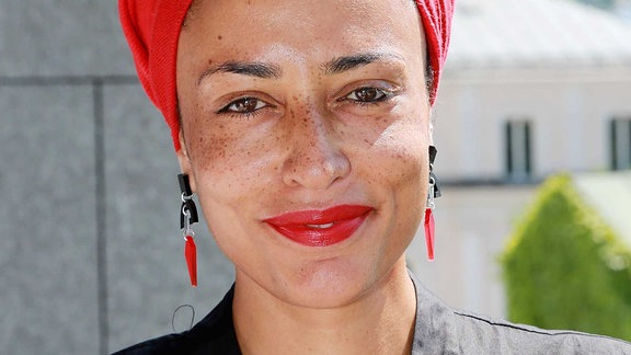 Eine Frau Anfang 40 mit Sommersprossen lächelt. Sie trägt ein rotes Tuch, passend zu Ohringen und Lippenstift.
