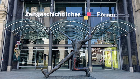 Der Eingang des Zeitgeschichtlichen Forums in Leipzig mit drei glasüberdachten Eingangswegen, davor steht die Plastik "Der Jahrhundertschritt"