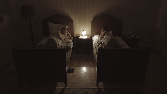 Ein Mann und eine Frau liegen in zwei einzelnen Betten in einem Schlafzimmer. Zwischen ihnen leuchtet eine Nachttischlampe, sie sind einander zugewandt.