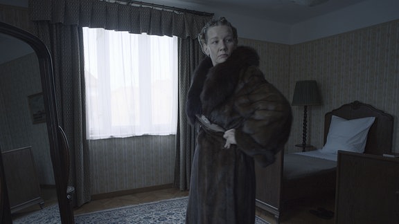 Eine Frau (Sandra Hüller) steht in einem Schlafzimmer und probiert vor dem Spiegel einen Pelzmantel an.