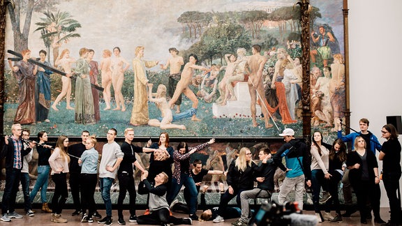 Die Auszubildenden des MDR stehen unter einem riesigen Gemälde im Leipziger Museum der bildenden Künst und stellen die Szenerie des Gemäldes nach.