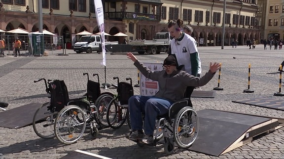 Eine Person schiebt eine andere Person in einem Rollstuhl über eine Rampe.