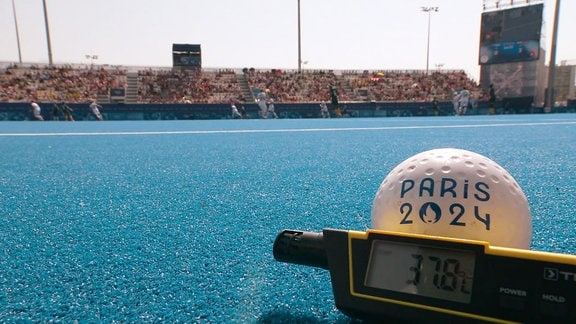 Ein Hockeyball liegt auf einem Hockeyfeld zusammen mit einem Thermometer, das 37,8 Grad Celsius anzeigt.