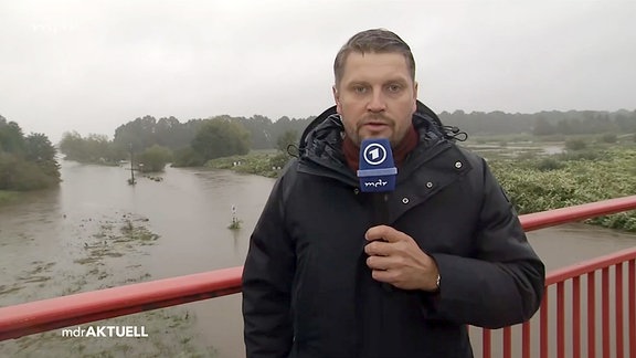 Ein Mann spricht in ein Mikrofon und steht auf einer Brücke die über einen Fluss führt, der über die Ufer tritt.