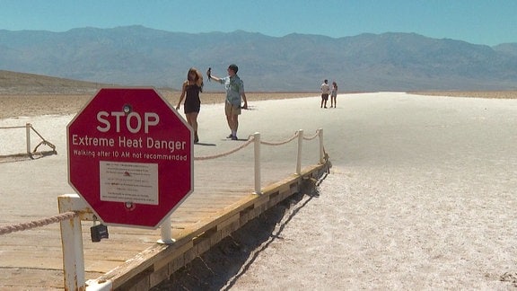 Warnschild vor Überhitzung vor einer Sandfläche, dahinter: Spaziergänger