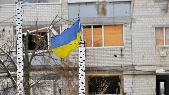 Die Nationalflagge der Ukraine steht vor einem zerstörten Gebäude.