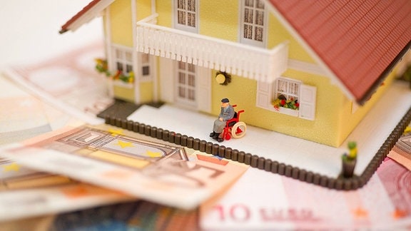 Eine Spielzeugfigur sitzt, im Rollstuhl, im Vorgarten eines Spielzeughauses, dass auf Geldscheinen steht.