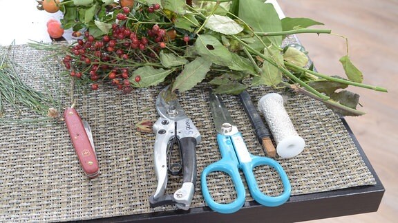 Auf einem Tisch liegen Schere, Gartenschere, Messer und verschiedene Zweige. 