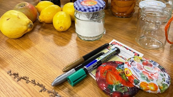 Schraubdeckel, Stifte, Eitketten, Obst und Marmeladengläser stehen auf einem Tisch.