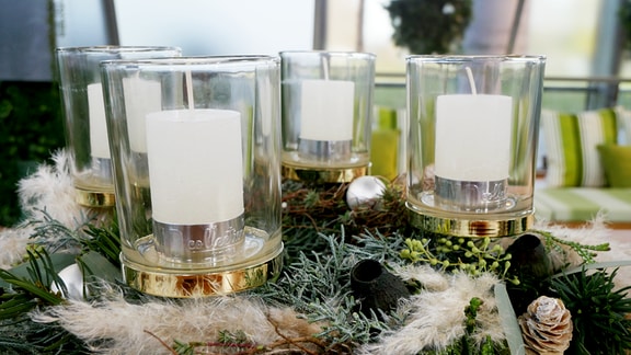 Ein Adventskranz mit Dekor um Kerzengläser drapiert. In den Gläsern stehen weiße Stumpenkerzen.