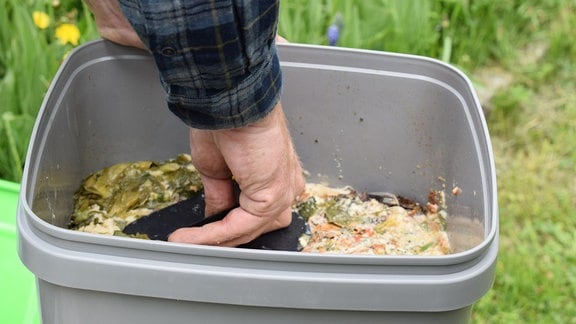 Eine Hand drückt mit einem Spatel aus schwarzem Kunststoff die Küchenabfälle in einem grauen Bokashi-Komposteimer fest