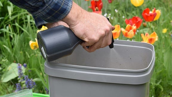 Eine Hand sprüht mit einer dunklen Sprühflasche Wasser in einen grauen Bokashi-Komposteimer