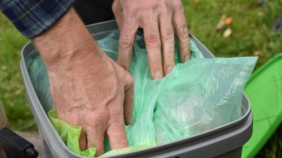 Hände drücken einen Müllbeutel mit Sand darin auf den Inhalt eines gefüllten grauen Bokashi-Komposteimers