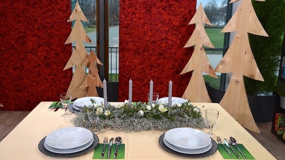 Auf einem gedeckten Tisch liegt eine längliche Weihnachtdeko mit grauen Kerzen, Christrosen und verschiedenen Kräutern und Tanne.