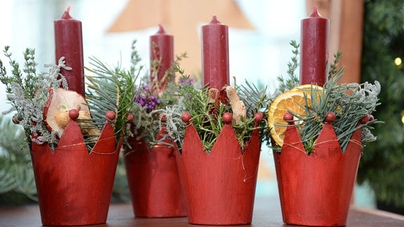In Roten Übertöpfen stecken rote Kerzen. Die Topfe sind mit Tanne, getrockneten Orangen und Äpfeln dekoriert.