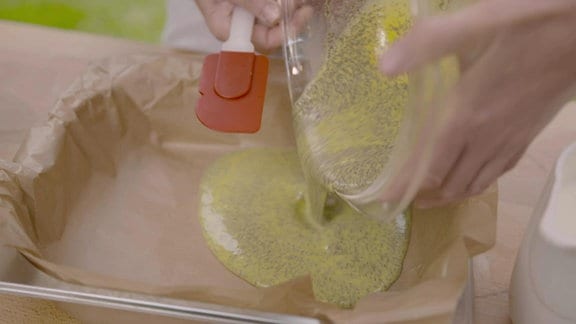 Aus einer Rührschüssel aus Kunststoff wird hellgelber Pfannkuchenteig mit gequetschten Mohnsamen darin in eine mit Backpapier ausgekleidete Backform gegossen