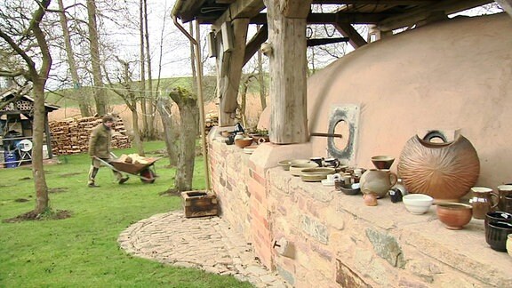 Ein Garten mit einem Ofen, in dem Keramik gebrannt wird. Im Hintergrund läuft jemand mit einer Schubkarre.