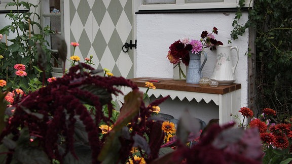 Vor einer Gartenlaube wächst roter Fuchsschwanz, auf einem Tisch stehen Vasen mit Blumen.