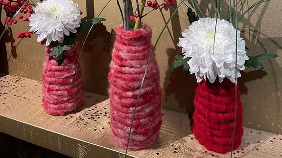Drei mit dicker, roter Wolle umwickelte Flaschen, in denen Blumen stecken.