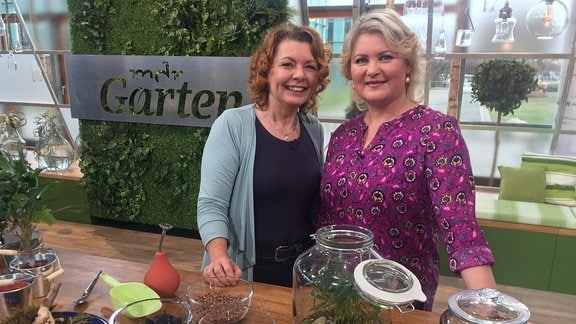 Gartenexpertin Brigitte Goss und Moderatorin Diana Fritzsche Grimmig stehen an einem Pflanztisch auf dem Pflanzen in Gläsern arrangiert wurden.