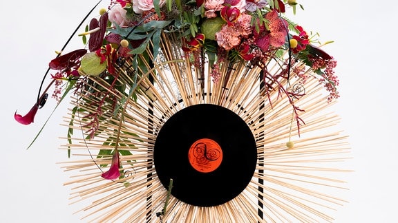 Ein Strahlenkranz aus Holzstäbchen, geschmückt mit Blumen, in der Mitte eine Schallplatte