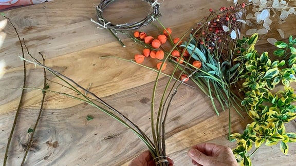 Zwei Hände binden Äste zusammen, auf einem Tisch liegen Pflanzenteile.