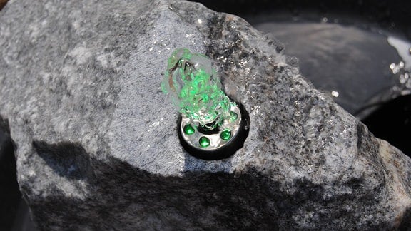 Aus einer Öffnung mit einem Schlauch in einem Stein quillt Wasser, das durch Leuchten in der Einfassung des Schlauches von unten grün angestrahlt wird