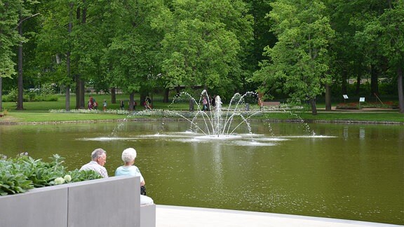 Zwei ältere Menschen sitzen an einem Teich. In der Mitte des Teiches ist ein Springbrunnen.