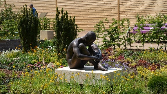 In der Mitte eines Beetes befindet sich eine bronzene Figur. Der junge sitzende nackte Mann hat die Arme um seine Beine geschlungen. Auf dem Beet wachsen viele verschiedene Blumen. 