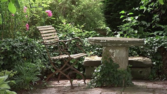 Ein alter Stuhl steht an einem alten Steintisch in einem Garten.