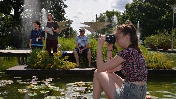 Ein Mädchensitzt an einem Teich und fotografiert. Im Hintergrund stehen Menschen.