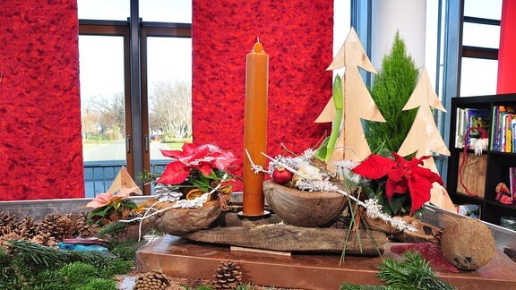 Auf einem Tisch mit Tannengrün und Tannenzweigen steht ein langes Holzstück auf der bepflanzte Kokusnusshälften und eine Kerze befestigt wurden