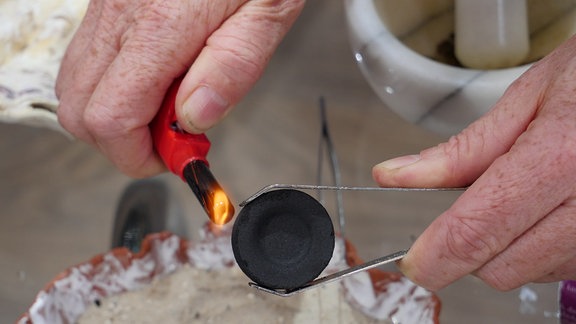 Räucherkohle anzünden - Eine Räucherkohle-Tablette wird mit einer Metallzange gehalten und mit einem Feuerzeug angezündet.