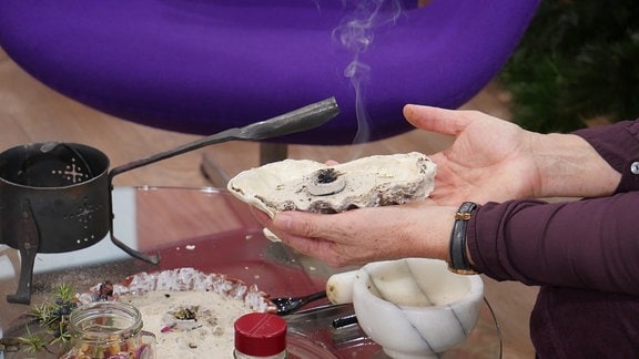 In einer großen Muschel mit Sand qualmt eine Räucherkohle-Tablette mit Kräutern. Zwei Hände halten die Muschel. Darunter ist ein Glastisch zusehen auf dem Räucherutensilien stehen.