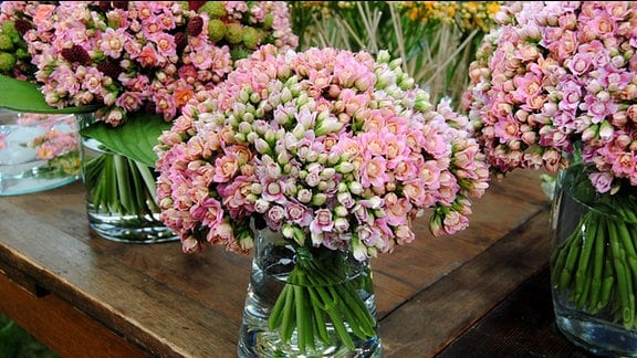 Drei gläserne Vasen mit dichten, kugelförmigen Blumensträußen mit rosa und weißen Blüten stehen auf einem Holztisch auf einem Rasenstück