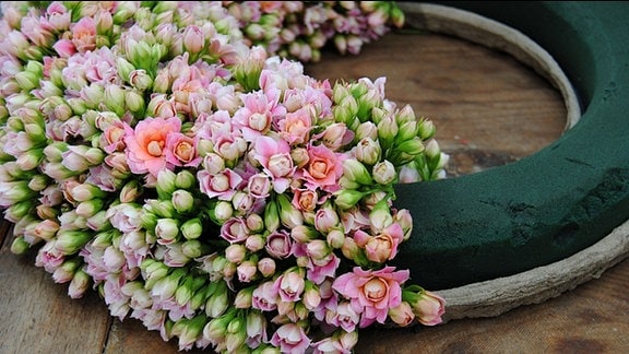 Auf einem großen Teil eines Ringes mit dunkelgrüner Steckmasse, der auf einem Holztisch liegt, wurden kleine rosa und weiße Blüten dicht beieinander befestigt