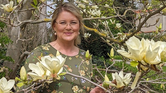 Eine Frau (Diana Fritzsche-Grimmig) steht an einem großen Baum voller gelber Blüten. Sie hält einen Ast vor ihre Brust